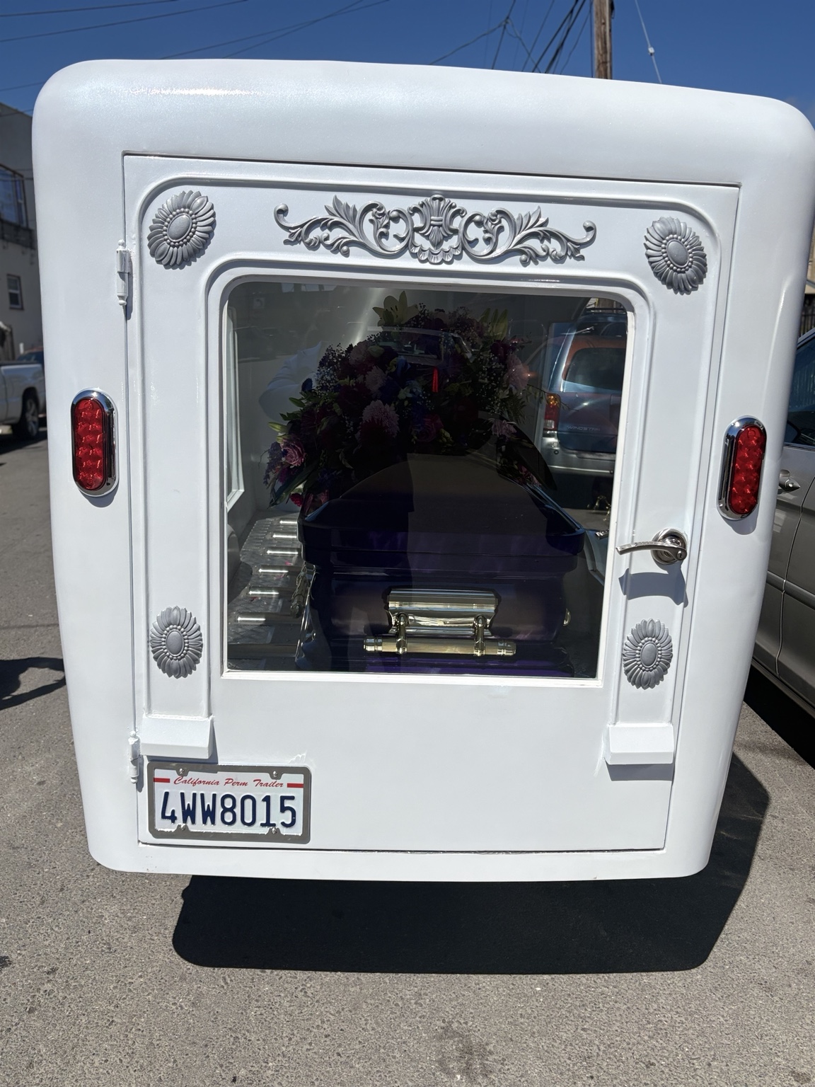 Rear view of white hearse trailer carrying a purple casket with floral arrangement